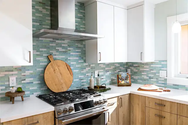 Kitchen with a sea-glass blue tile backsplash, white upper cabinets, light wood lower cabinets and a stainless range